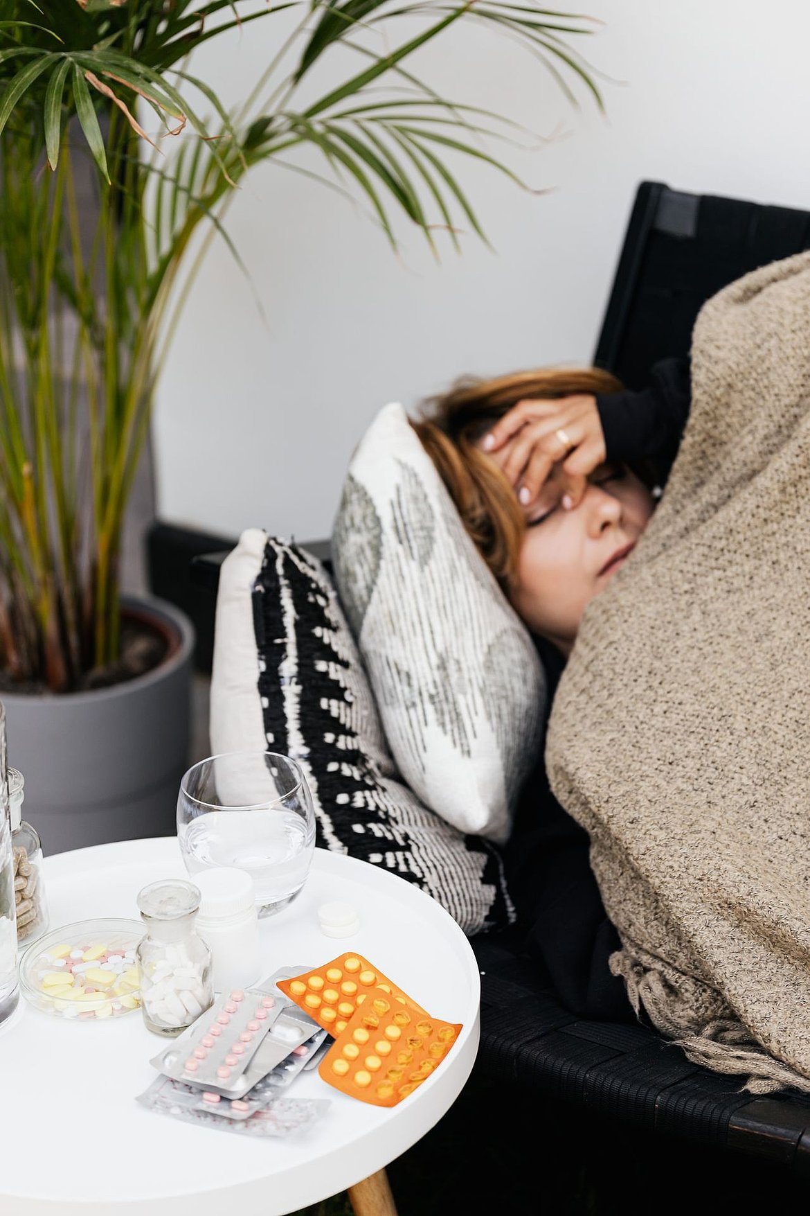 Frau liegt auf einem Sofa, mit einer Decke bedeckt, fühlt sich unwohl, mit Pillen auf einem Tisch in der Nähe.