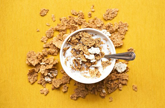 Das Foto zeigt eine Schüssel mit Cornflakes von oben fotografiert. Die Schüssel steht auf einer gelben Tischdecke. Rund um die Schüssel liegen Krümel der Flakes auf der Tischdecke.
