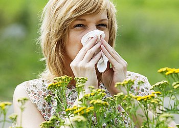 Junge Frau mit Heuschnupfen und Allergie steht in Frühlingswiese und putzt sich die Nase.