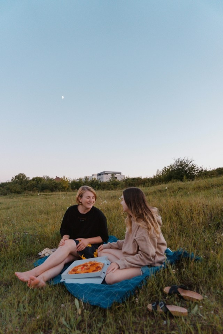 Das Foto zeigt zwei junge Frauen. Sie sitzen in der Dämmerung auf einer Wiese und unterhalten sich.
