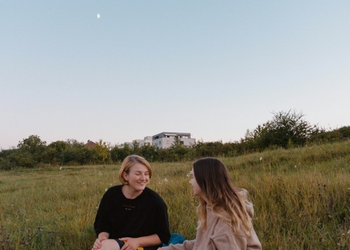 Das Foto zeigt zwei junge Frauen. Sie sitzen in der Dämmerung auf einer Wiese und unterhalten sich. 