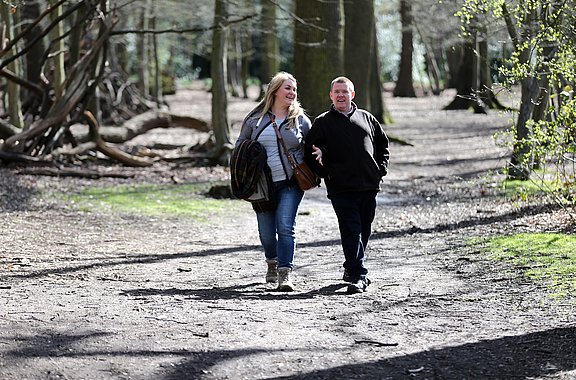Das Foto zeigt einen Mann und eine Frau beim Spaziergang in einem Park. Die Sonne scheint, die Bäume haben erstes Grün an den Ästen.