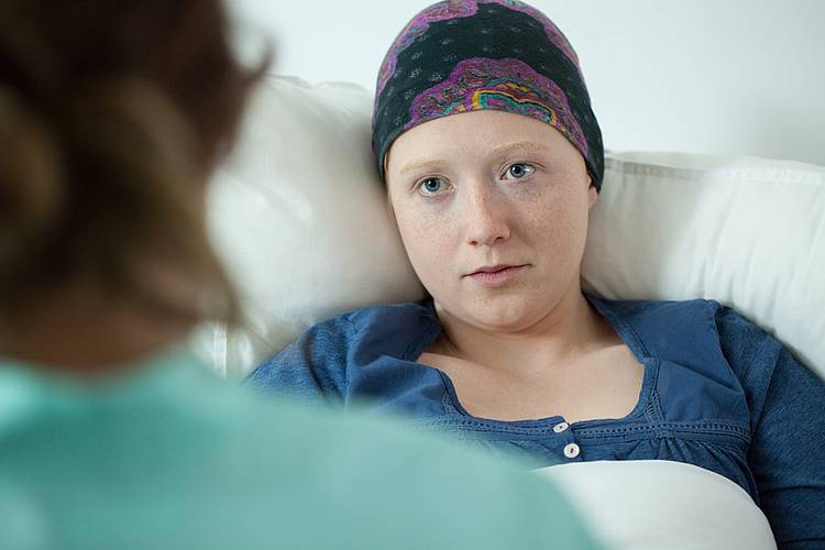 Junge Krebspatientin im Krankenbett ist im Gespräch mit einer Krankenschwester oder Ärztin.