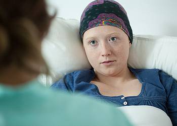 Junge Krebspatientin im Krankenbett ist im Gespräch mit einer Krankenschwester oder Ärztin.