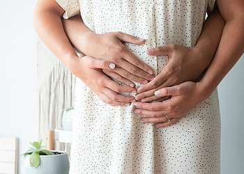 Das Foto zeigt im Bildausschnitt den Bauch einer schwangeren Frau. Vater und Mutter haben schützen die Hände über den Bauch gelegt.