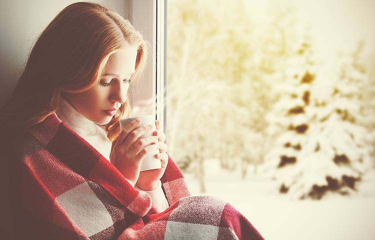 Junge Frau wärmt sich an einem Wintertag mit Tee in der Hand und einer Decke um die Schultern. Sie friert, weil sie an Blutarmut leidet.