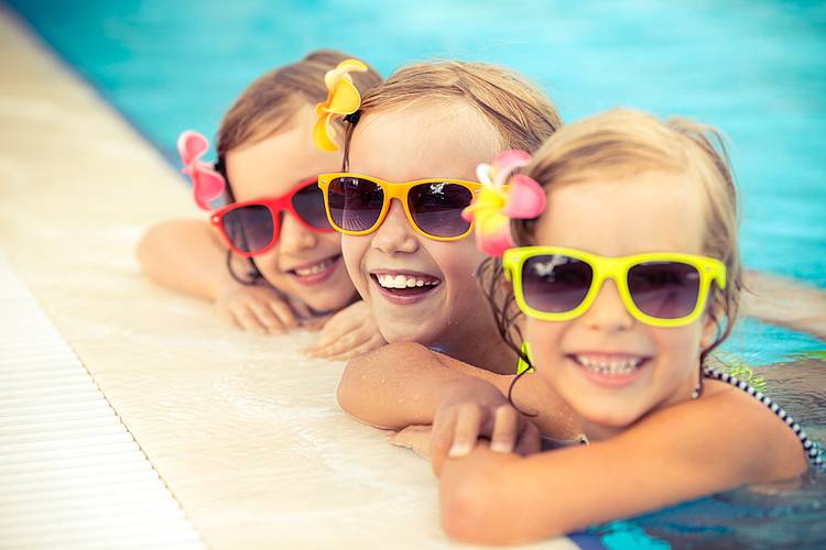 Drei kleine Mädchen mit Sonnenbrille halten sich am Schwimmbeckenrand im Freibad fest.