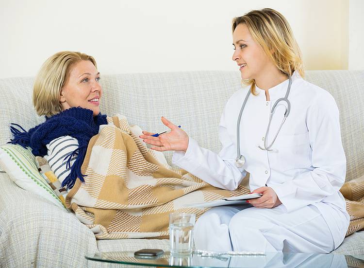 Junge Ärztin erklärt beim Hausbesuch etwas einer kranken Patienten auf einem Sofa.