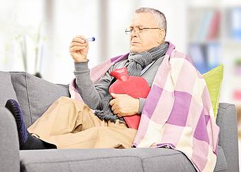 Kranker älterer Mann liegt mit Fieberthermometer und Wärmflasche auf dem Sofa.