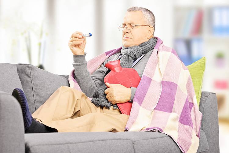 Kranker älterer Mann liegt mit Fieberthermometer und Wärmflasche auf dem Sofa.